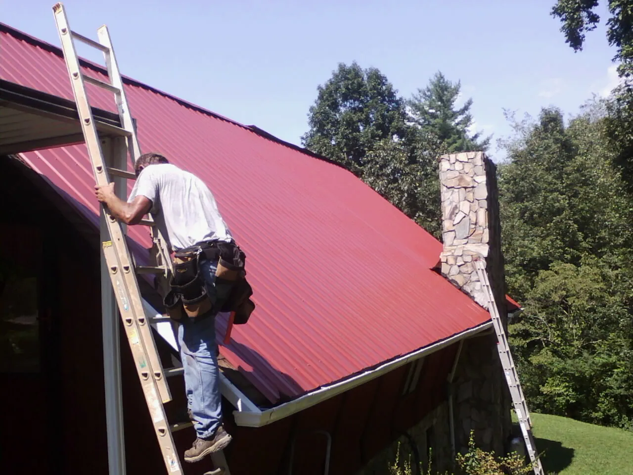 Roofer on a ladder beside a red metal roof and stone chimney — Soffit & Fascia Repair by Roofing Brothers in Amagansett